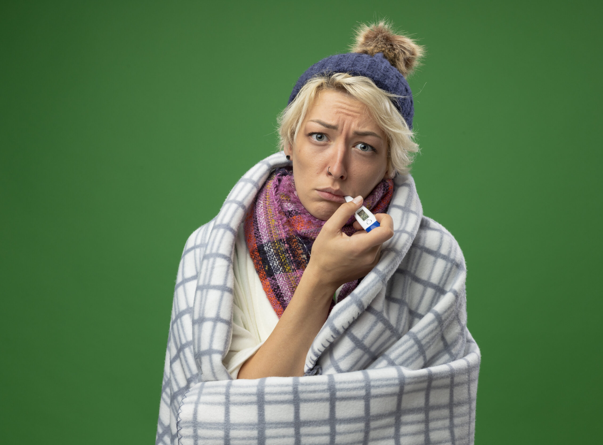 Serganti moteris su žiemine kepure, šaliku ir pledu, matuojasi temperatūrą termometru burnoje